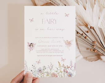 Może przedstawiać: Białe zaproszenie na baby shower z napisem a little FAIRY is on her way, ozdobione akwarelowymi ilustracjami dwóch wróżek, motyli oraz kwiatową bordiurą u dołu w odcieniach różu, fioletu i zieleni.