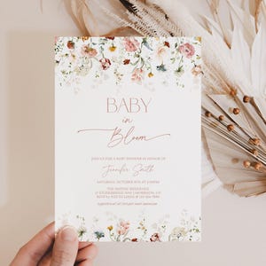 Op de afbeelding: Een uitnodiging voor een babyshower met bloemaccenten. De uitnodiging bevat de woorden "BABY in Bloom" in een sierlijk handschrift. Bloemsierkunst in roze, wit en groen versieren de boven- en onderranden.