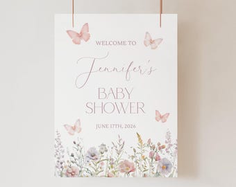 May include: A white rectangular baby shower welcome sign featuring pink watercolor butterflies and a bottom border of wildflowers. The text reads: WELCOME TO Jennifer's BABY SHOWER JUNE 17TH, 2026. The sign is suspended by thin copper-colored metal rods.