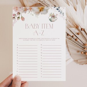 May include: A baby shower game card with floral watercolor accents. The card features the text "BABY ITEM A-Z" in a mauve font, with lines for writing baby item names next to each letter of the alphabet. The top of the card is decorated with watercolor flowers.