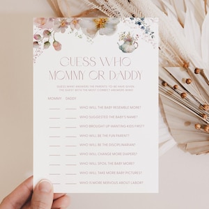 May include: A baby shower game card with the text "Guess Who Mommy or Daddy". The card has a floral design at the top and prompts for answers to questions about the parents-to-be. The card is held by a hand.