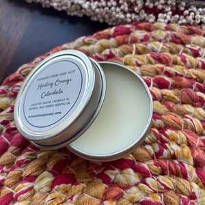 May include: A small tin of Healing Orange Calendula hand salve, with a white label and black text. The label reads "Cardman's Friend Hand Salve Healing Orange Calendula Grass-fed Tallow, Calendula Oil, Beeswax, Wild Orange Essential Oil aroundthespinney.com". The tin is sitting on a colorful woven mat.