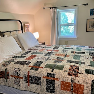 May include: A bed with a black metal frame is covered with a quilt featuring a geometric pattern in shades of beige, black, green, and orange. White pillows and a white bed skirt complete the bedding ensemble. The room has a window with white curtains.