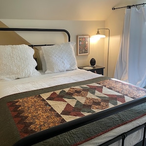 May include: A bed with a black metal frame, white bedding, and a patterned quilt. The quilt features geometric shapes in shades of brown, green, and red. Two white pillows are on the bed. A floor lamp and a framed picture are visible.