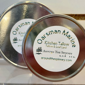 May include: Two silver metal tins with white labels. The larger tin has the text "Oarsman" partially visible. The smaller tin has the text "Oarsman Marine" and the text "Kitchen Tallow Tallow & Leaf Lard" and the text "AROUND THE SPINNEY and sea aroundthespinney.com".