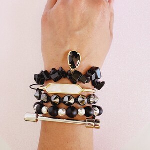 May include: A stack of bracelets featuring black and silver beads, a white rectangular charm, and a black gemstone pendant. The bracelets are worn on a person's wrist.