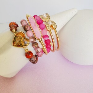 May include: A set of five bracelets featuring pink and brown gemstone beads, a gold heart charm, and a gold bangle with a pearl and rhinestone design.