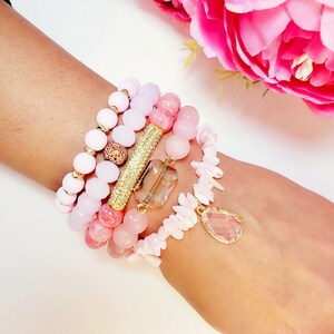 May include: A set of pink and white beaded bracelets with gold accents. The bracelets feature various shapes and sizes of beads, including round, oval, and rectangular. The gold accents include a bar with rhinestones and a teardrop pendant.