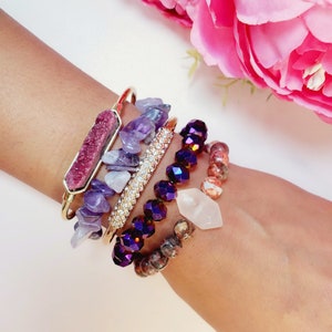May include: A set of five bracelets featuring various gemstone and crystal accents. The bracelets include a gold bangle with a pink crystal, a gold bangle with purple and white stones, a gold bangle with clear crystals, a purple beaded bracelet, and a brown and pink beaded bracelet.
