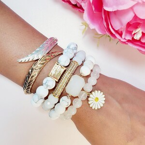 May include: A stack of white and gold bracelets with a daisy charm. The bracelets are made of white beads and gold accents. The daisy charm is white with a yellow center.
