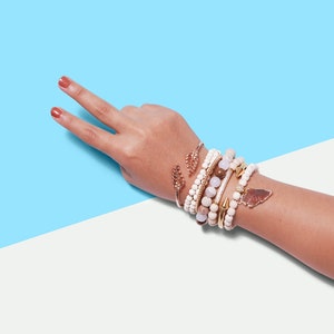 Op de afbeelding: Een pols is versierd met een selectie armbanden in diverse stijlen en kleuren. De armbanden hebben kralen-, gouden en stenen ontwerpen. Een gouden manchetarmband met bladaccenten is ook aanwezig. De achtergrond is verdeeld tussen lichtblauw en wit.