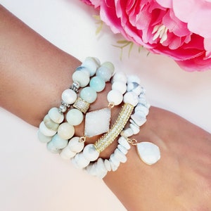 May include: A stack of bracelets featuring white and light green stones, a gold bracelet with sparkling stones, and a white teardrop pendant.