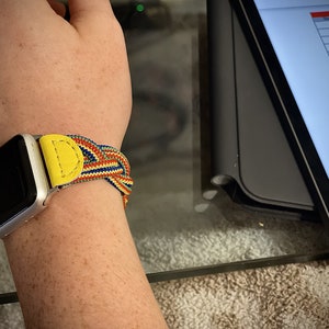 Puede incluir: Reloj inteligente plateado con una colorida correa trenzada. La correa presenta detalles de cuero amarillo y un patr&oacute;n tejido de hilos rojos, azules y amarillos. El reloj est&aacute; en una mu&ntilde;eca.