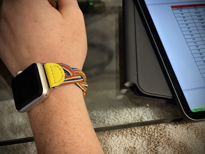 Puede incluir: Reloj inteligente plateado con una colorida correa trenzada. La correa presenta detalles de cuero amarillo y un patr&oacute;n tejido de hilos rojos, azules y amarillos. El reloj est&aacute; en una mu&ntilde;eca.