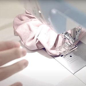 Pu&ograve; includere: Primo piano di un tessuto rosa che viene cucito su una macchina da cucire bianca. Il tessuto viene inserito nell'ago e il piedino della macchina preme su di esso.