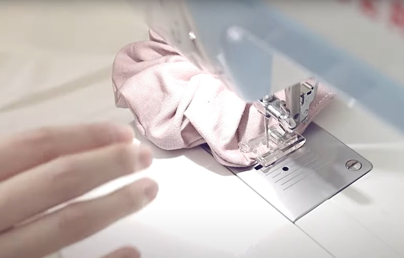 Pu&ograve; includere: Primo piano di un tessuto rosa che viene cucito su una macchina da cucire bianca. Il tessuto viene inserito nell'ago e il piedino della macchina preme su di esso.