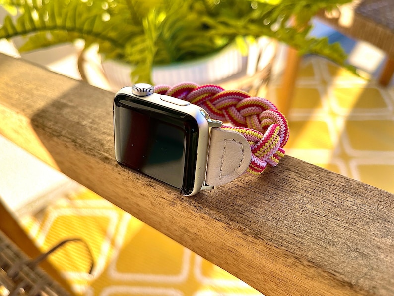 Puede incluir: Reloj inteligente plateado con una correa trenzada en tonos rosa, amarillo y rojo. La correa del reloj est&aacute; hecha de un material tejido similar a una cuerda y est&aacute; sujeta al reloj con una correa de cuero de color claro. El reloj est&aacute; sobre una superficie de madera.