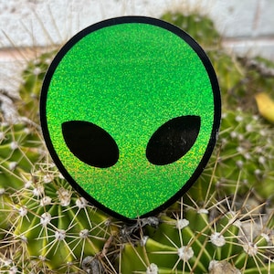 May include: Green glitter alien head sticker with black outline and eyes. The sticker is on a green cactus.