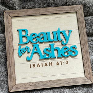 May include: Wooden framed sign with the words "Beauty for Ashes" in turquoise, above the text "Isaiah 61:3". The sign has a light wood background with horizontal lines and a brown wooden frame.