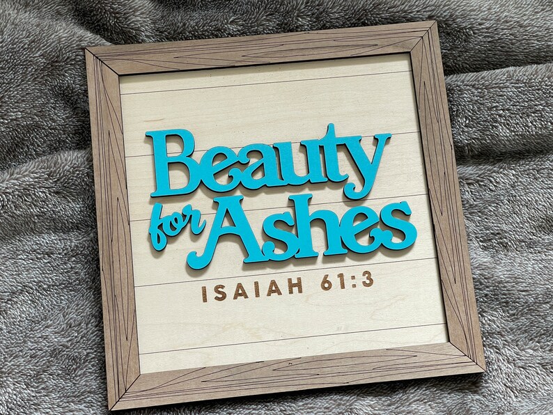 May include: Wooden framed sign with the words "Beauty for Ashes" in turquoise, above the text "Isaiah 61:3". The sign has a light wood background with horizontal lines and a brown wooden frame.