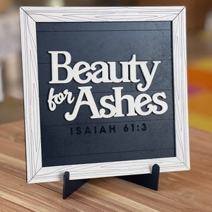 May include: A framed wooden sign with the words "Beauty for Ashes" in white, set against a dark blue background. The frame is white with a wood grain design. The text "Isaiah 61:3" is below the main text. The sign is displayed on a wooden surface.