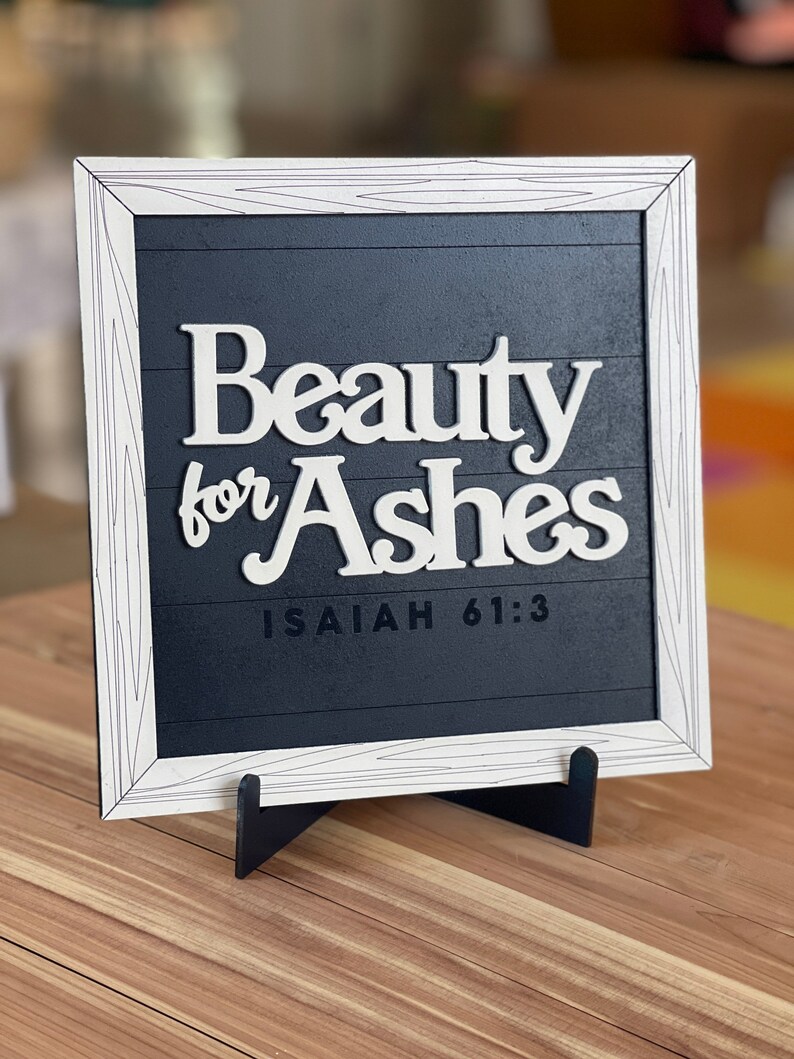 May include: A framed wooden sign with the words "Beauty for Ashes" in white, set against a dark blue background. The frame is white with a wood grain design. The text "Isaiah 61:3" is below the main text. The sign is displayed on a wooden surface.