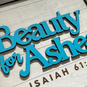 May include: Wooden sign with the words "Beauty for Ashes" in light blue letters with black outlines. The words are layered on a light wood background with the text "Isaiah 61:3" in brown. The sign is framed in a light brown wood.