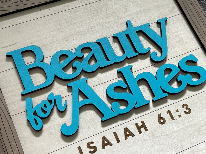 May include: Wooden sign with the words "Beauty for Ashes" in light blue letters with black outlines. The words are layered on a light wood background with the text "Isaiah 61:3" in brown. The sign is framed in a light brown wood.