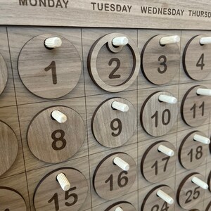 May include: Wooden perpetual calendar with engraved numbers and days of the week. The calendar features circular wooden date markers with white pegs. The days of the week are printed across the top in a sans-serif font.