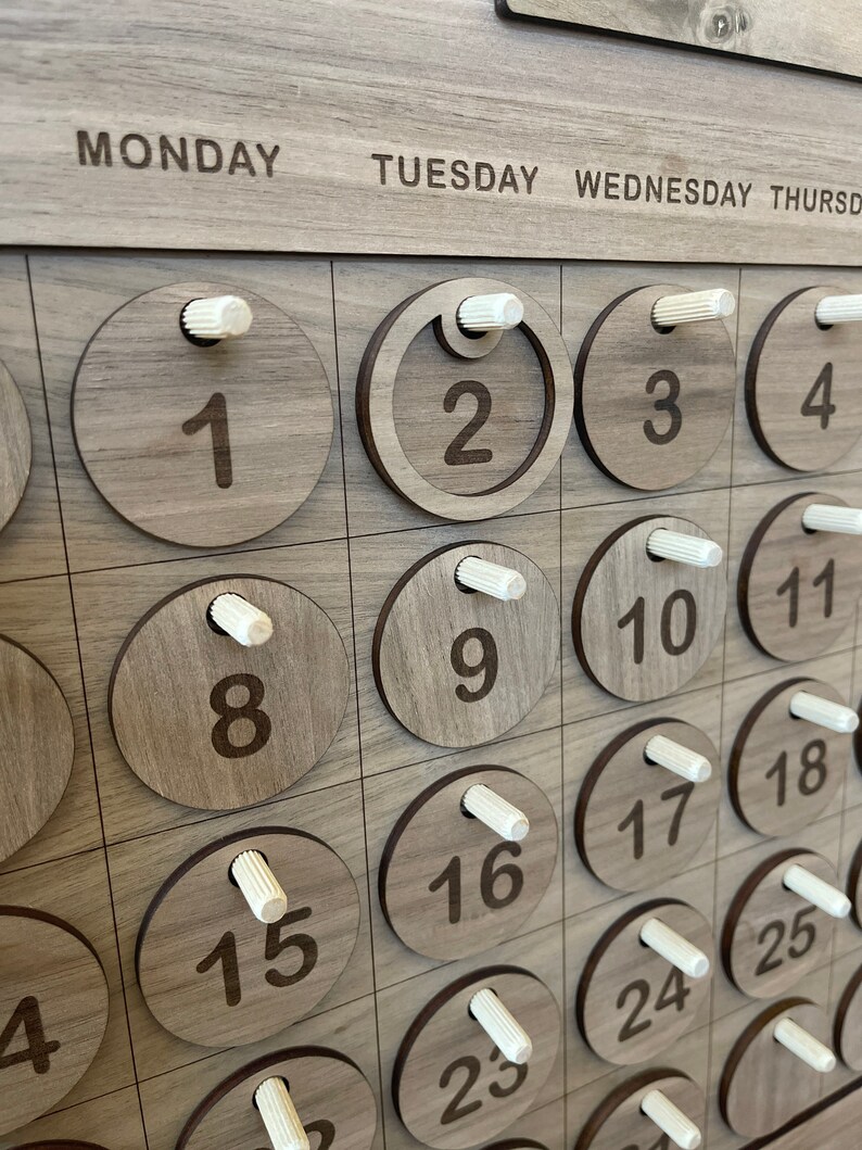 May include: Wooden perpetual calendar with engraved numbers and days of the week. The calendar features circular wooden date markers with white pegs. The days of the week are printed across the top in a sans-serif font.