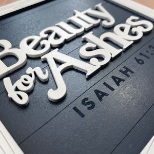 May include: A framed sign with the words "Beauty for Ashes" in raised white letters on a dark blue background. The text "Isaiah 61:3" is printed below. The frame is white.