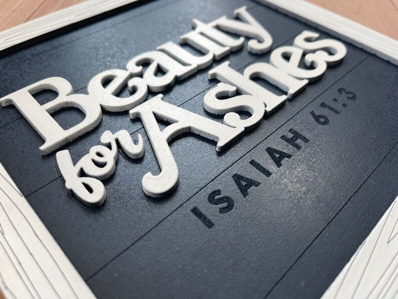 May include: A framed sign with the words "Beauty for Ashes" in raised white letters on a dark blue background. The text "Isaiah 61:3" is printed below. The frame is white.