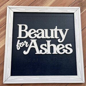 May include: White framed sign with the words "Beauty for Ashes" in a cursive font. The sign has a dark blue background with horizontal lines and the text "Isaiah 61:3" at the bottom. The frame is white with a wood grain design.