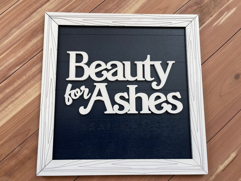 May include: White framed sign with the words "Beauty for Ashes" in a cursive font. The sign has a dark blue background with horizontal lines and the text "Isaiah 61:3" at the bottom. The frame is white with a wood grain design.