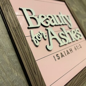 May include: A framed wooden sign with the words "Beauty for Ashes" in white, raised lettering. The background is a light pink color with horizontal lines. The text "Isaiah 61:3" is below the main text.
