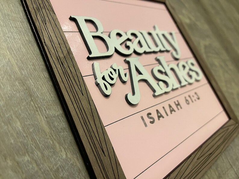 May include: A framed wooden sign with the words "Beauty for Ashes" in white, raised lettering. The background is a light pink color with horizontal lines. The text "Isaiah 61:3" is below the main text.