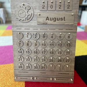 May include: Wooden perpetual calendar with the month of August displayed. The calendar features a sun and ball design, the year 2022, and the days of the week. The calendar has wooden pegs to mark the date.