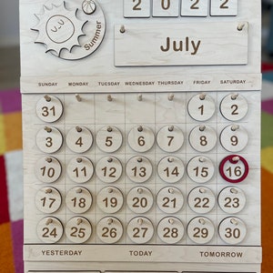 May include: Wooden July calendar with a sun and ball graphic, featuring the year 2022. The calendar displays the days of the week and numbered circles for the month. The 16th is highlighted in red. A wooden stand supports the calendar.