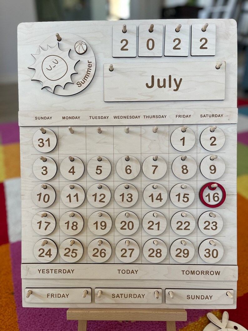 May include: Wooden July calendar with a sun and ball graphic, featuring the year 2022. The calendar displays the days of the week and numbered circles for the month. The 16th is highlighted in red. A wooden stand supports the calendar.