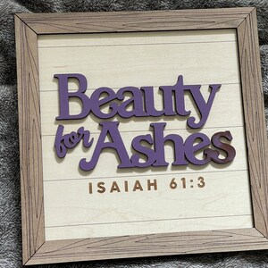May include: Wooden framed sign with the words "Beauty for Ashes" in purple, over a light wood background. The text "Isaiah 61:3" is printed in brown. The frame is a light brown color.