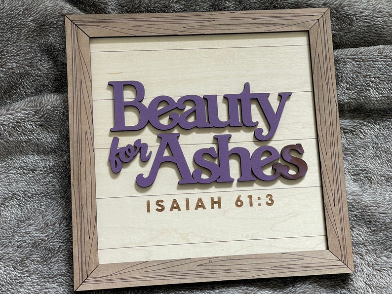 May include: Wooden framed sign with the words "Beauty for Ashes" in purple, over a light wood background. The text "Isaiah 61:3" is printed in brown. The frame is a light brown color.