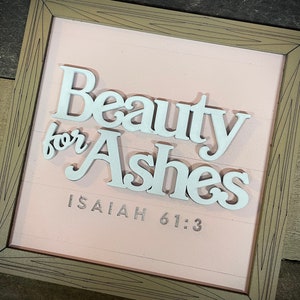 May include: A framed sign with the words "Beauty for Ashes" in white, raised lettering. The background is a light pink color with horizontal lines. The text "Isaiah 61:3" is printed below the main text. The frame is a light brown color.