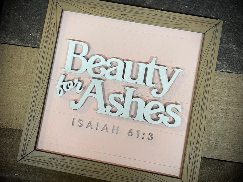 May include: A framed sign with the words "Beauty for Ashes" in white, raised lettering. The background is a light pink color with horizontal lines. The text "Isaiah 61:3" is printed below the main text. The frame is a light brown color.