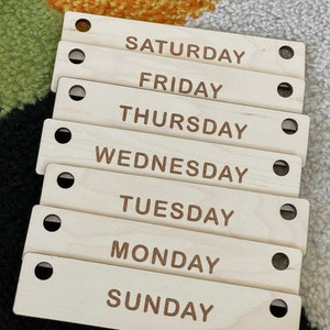 May include: Seven wooden rectangular tags, each labeled with a day of the week: Sunday, Monday, Tuesday, Wednesday, Thursday, Friday, and Saturday. Each tag has a hole on each end. The text is in a brown font.