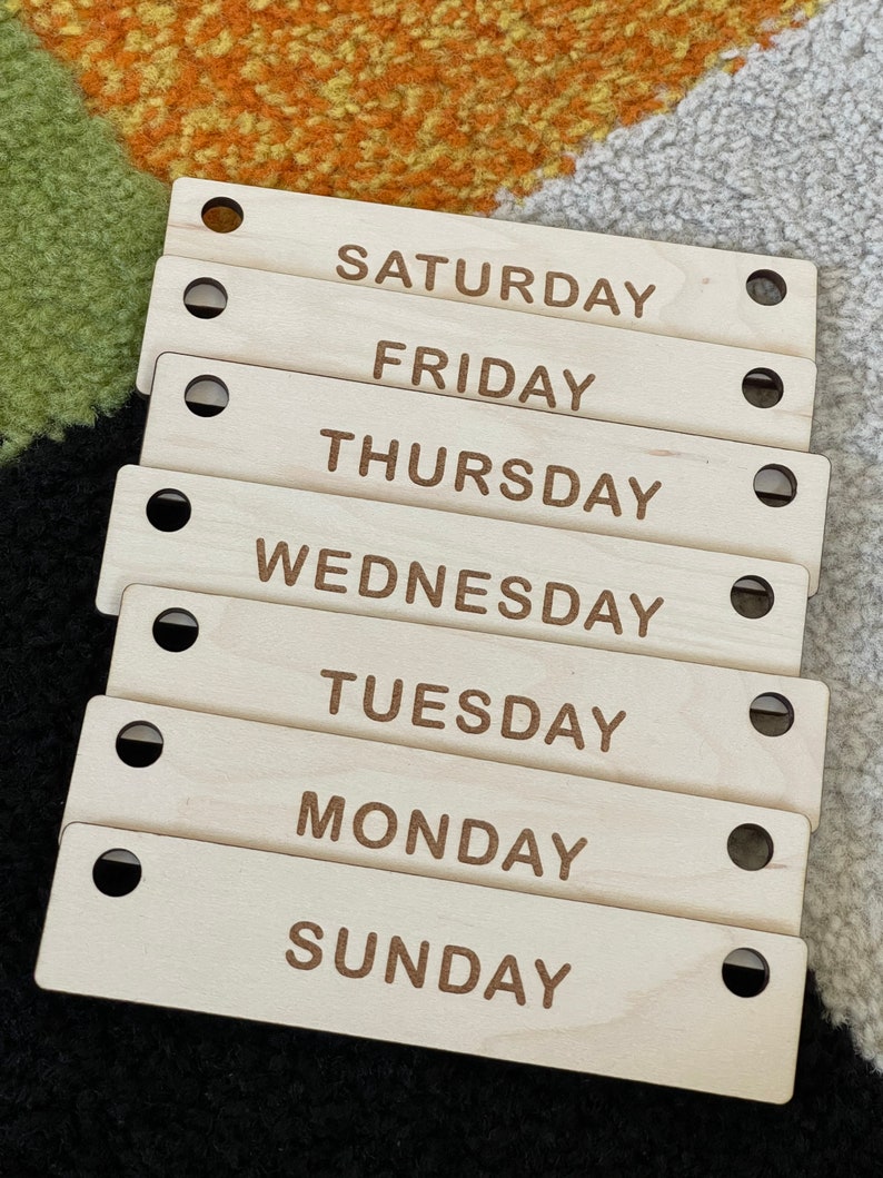 May include: Seven wooden rectangular tags, each labeled with a day of the week: Sunday, Monday, Tuesday, Wednesday, Thursday, Friday, and Saturday. Each tag has a hole on each end. The text is in a brown font.