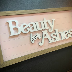 May include: A rectangular wooden sign with the words "Beauty for Ashes" in white, raised lettering. The background is a light pink, horizontal plank design, framed by a light brown wood-grain border. The sign is angled.