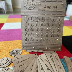 May include: Wooden perpetual calendar with interchangeable month and date pieces. The calendar includes a sun and ball graphic, the month of August, and the year 2022. Additional wooden pieces display the months and seasons.