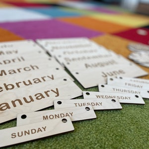 May include: Wooden calendar pieces with months and days of the week engraved in a simple font. The months are on larger rectangular pieces, and the days are on smaller, individual pieces. The wood has a natural, light color.