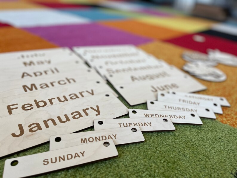 May include: Wooden calendar pieces with months and days of the week engraved in a simple font. The months are on larger rectangular pieces, and the days are on smaller, individual pieces. The wood has a natural, light color.