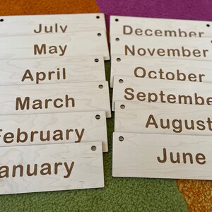 May include: Wooden month name plaques. Each plaque is rectangular with rounded corners and two small holes. The months are printed in a brown font. The months are: July, May, April, March, February, January, December, November, October, September, August, and June.